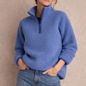 May include: A periwinkle blue fleece pullover with a high collar and a quarter-zip closure. The long-sleeved top has a soft, textured appearance and is paired with blue jeans. The collar and cuffs are trimmed with a darker blue fabric.