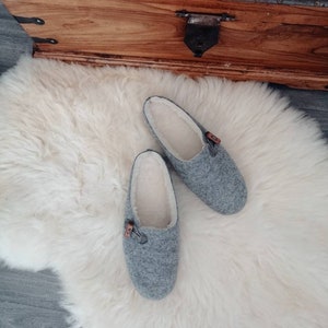 May include: A pair of grey wool slippers with a white fleece lining. The slippers have a brown button detail on the top.