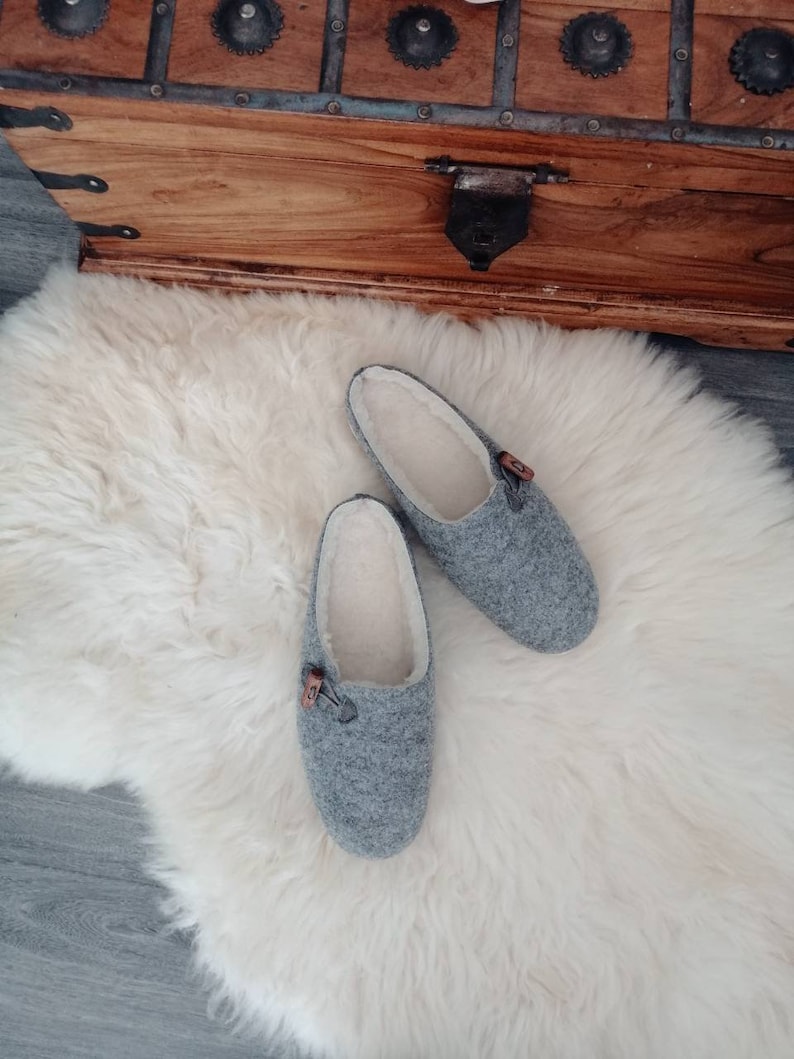 May include: A pair of grey wool slippers with a white fleece lining. The slippers have a brown button detail on the top.