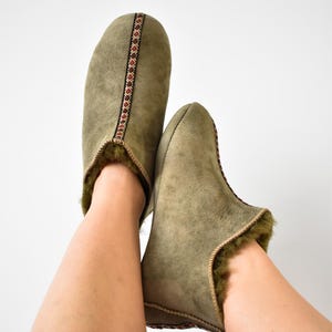 Könnte beinhalten: Ein Paar grüne Slipper aus Wildleder mit einem braunen und roten Muster am Rand. Die Slipper sind mit Fell gefüttert und haben eine geschlossene Spitze.