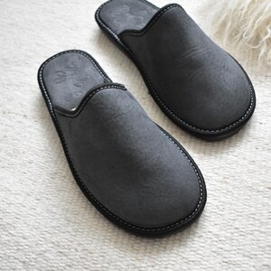 May include: A pair of gray suede slippers with black trim and white stitching. The slippers have a rounded toe and a simple design. The background is a white, textured surface.