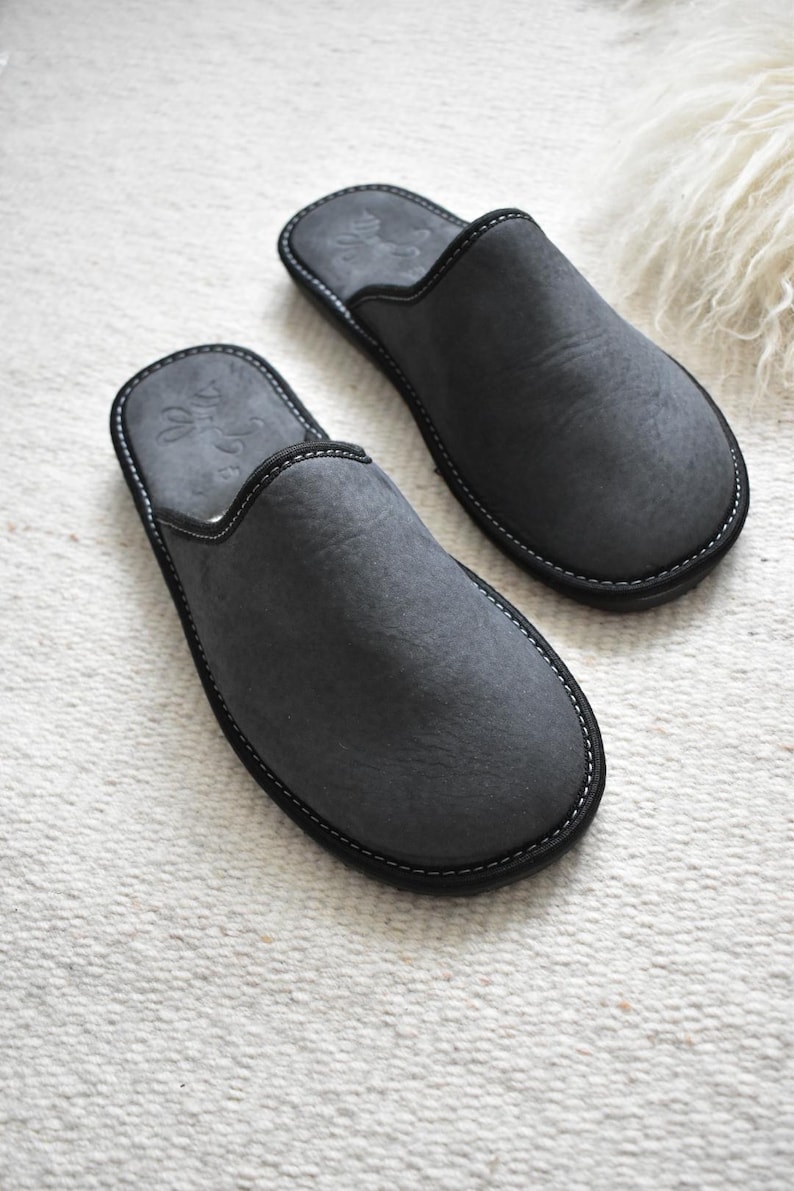 May include: A pair of gray suede slippers with black trim and white stitching. The slippers have a rounded toe and a simple design. The background is a white, textured surface.