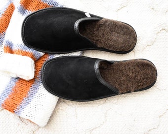 May include: A pair of black suede slippers with brown faux fur lining. The slippers are on a white fluffy rug.