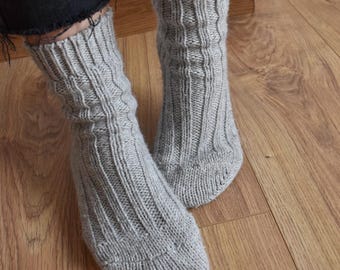 May include: A pair of grey knitted socks with a ribbed pattern. The socks are worn on a person's feet and are shown against a wooden floor.