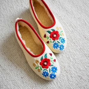 May include: A pair of white felt slippers with red and blue embroidered flowers. The slippers have a red trim around the edges.