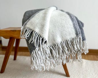 May include: A folded gray, white, and light gray fringed throw blanket draped over a small wooden stool. The blanket has a soft, textured appearance and is made of a thick material. The fringe is a light gray color.