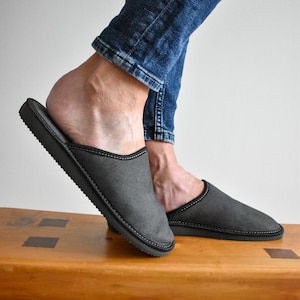 May include: Gray slip-on slippers with black trim and stitching. The slippers are on a wooden bench, with a person wearing blue jeans. The slippers appear to be made of a soft material.