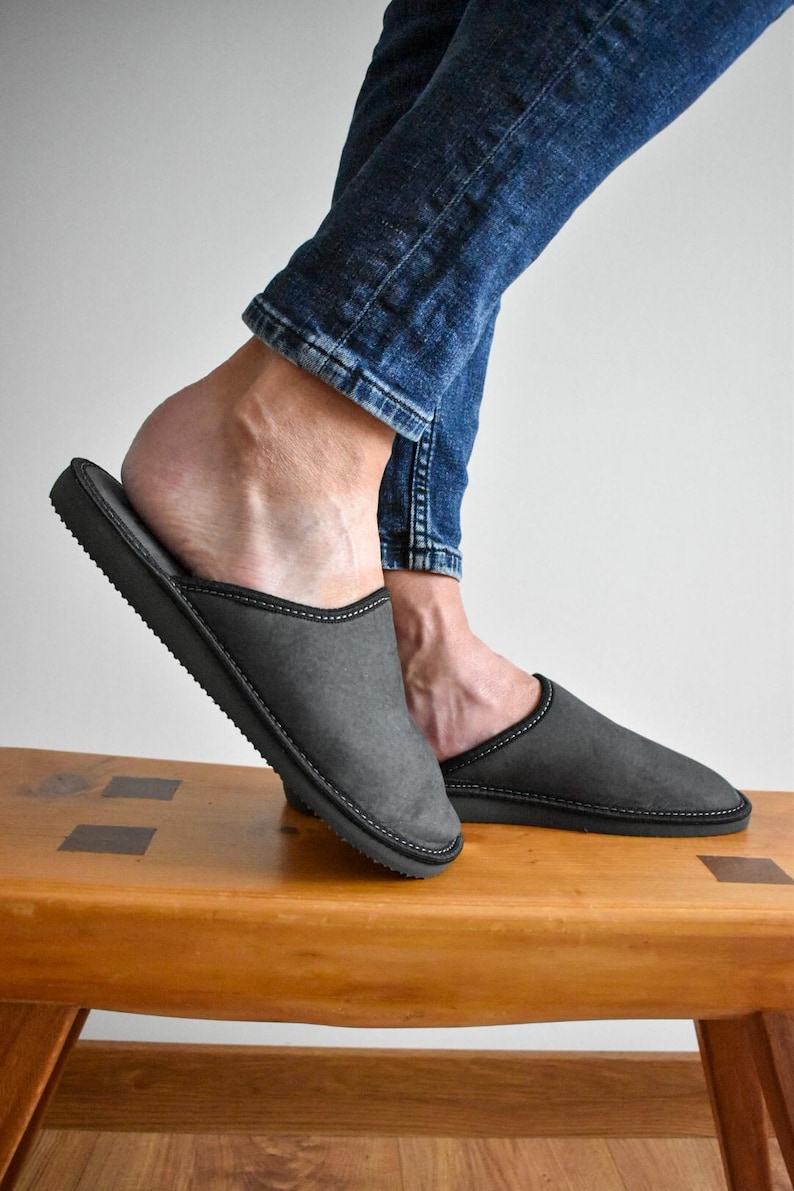 May include: Gray slip-on slippers with black trim and stitching. The slippers are on a wooden bench, with a person wearing blue jeans. The slippers appear to be made of a soft material.