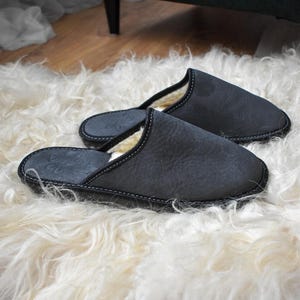 May include: A pair of dark gray leather slippers with white stitching and a soft, cream-colored lining. The slippers are resting on a fluffy, white sheepskin rug. The slippers have a simple, classic design.