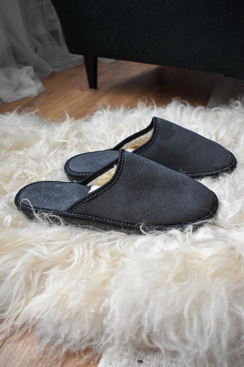 May include: A pair of dark gray leather slippers with white stitching and a soft, cream-colored lining. The slippers are resting on a fluffy, white sheepskin rug. The slippers have a simple, classic design.