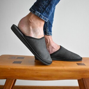 May include: Gray slippers with black trim are displayed on a wooden bench. The slippers have a simple design and a dark gray color. The image shows the slippers from a side view, highlighting their shape and construction.