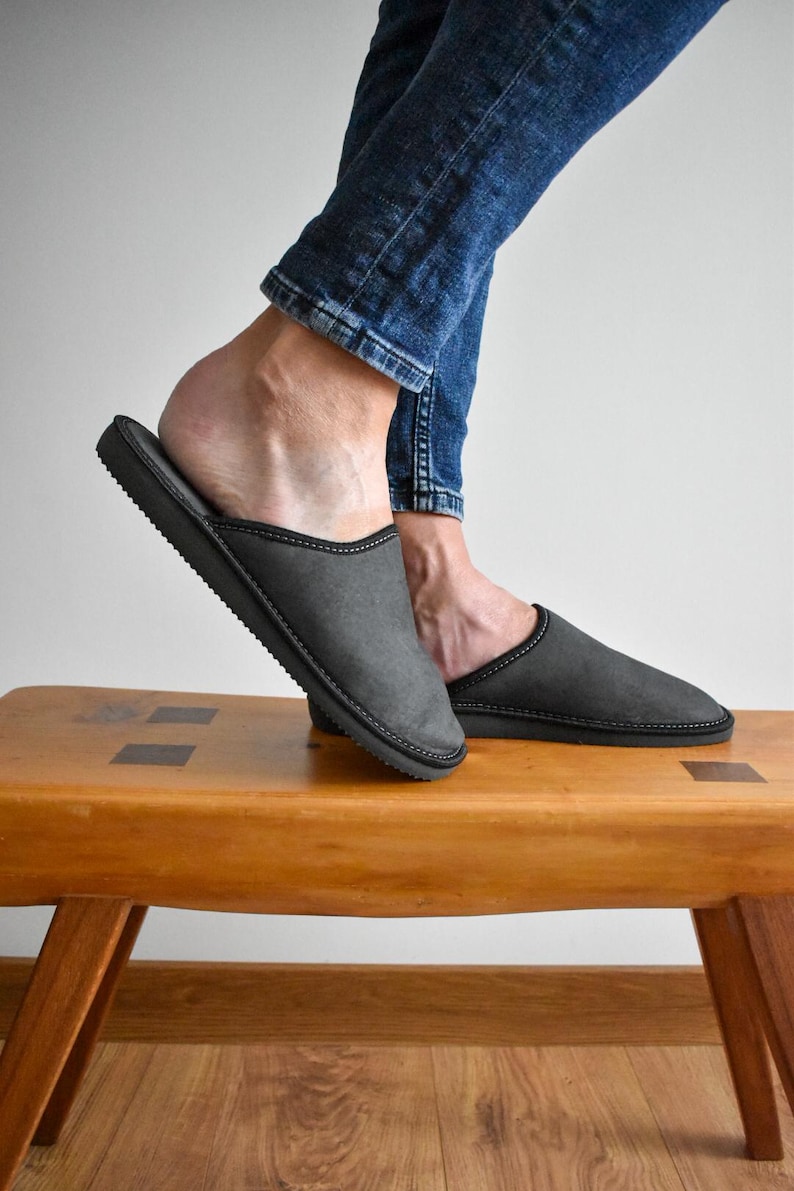 May include: Gray slippers with black trim are displayed on a wooden bench. The slippers have a simple design and a dark gray color. The image shows the slippers from a side view, highlighting their shape and construction.