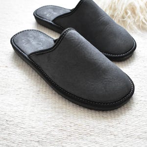 May include: A pair of charcoal gray suede slippers with a black sole and white stitching. The slippers have a rounded toe and a slip-on design. The interior features a subtle embossed design. The slippers are designed for indoor use.
