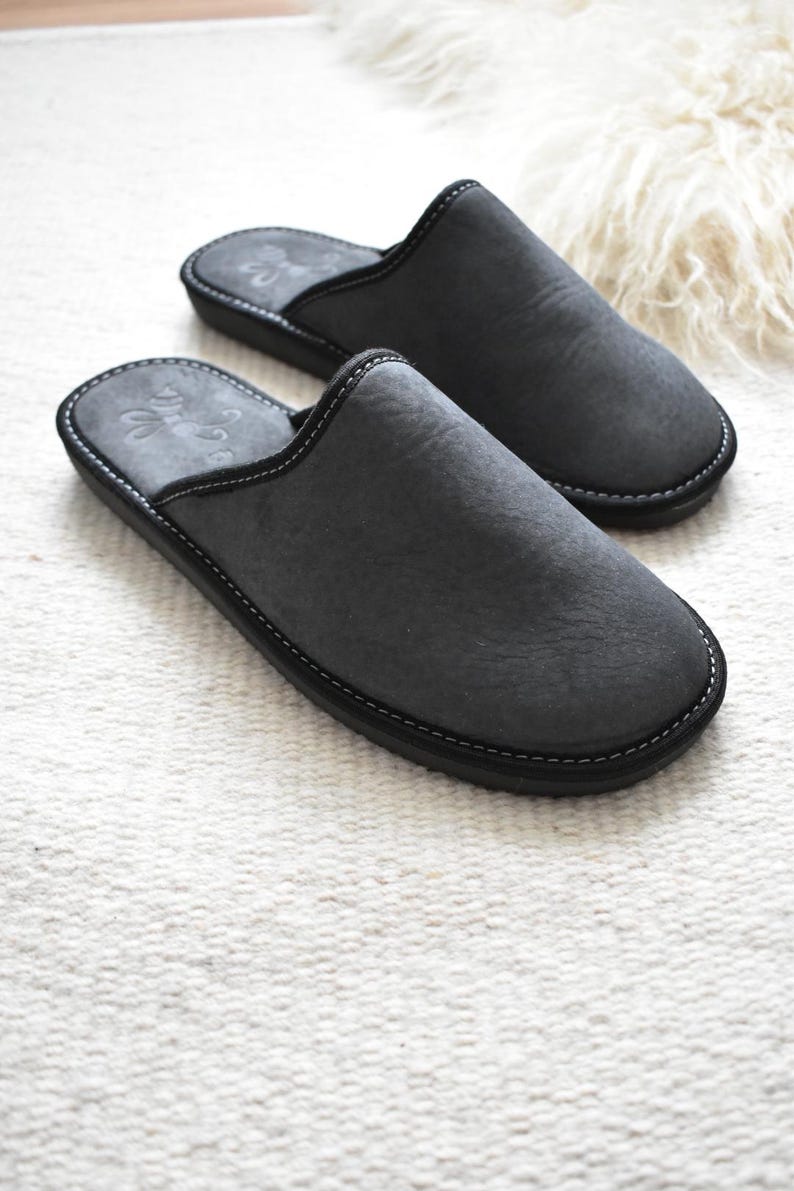 May include: A pair of charcoal gray suede slippers with a black sole and white stitching. The slippers have a rounded toe and a slip-on design. The interior features a subtle embossed design. The slippers are designed for indoor use.