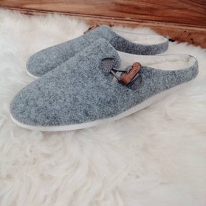 May include: A pair of grey wool slippers with a wooden buckle detail. The slippers have a white sole and a soft, fuzzy lining.