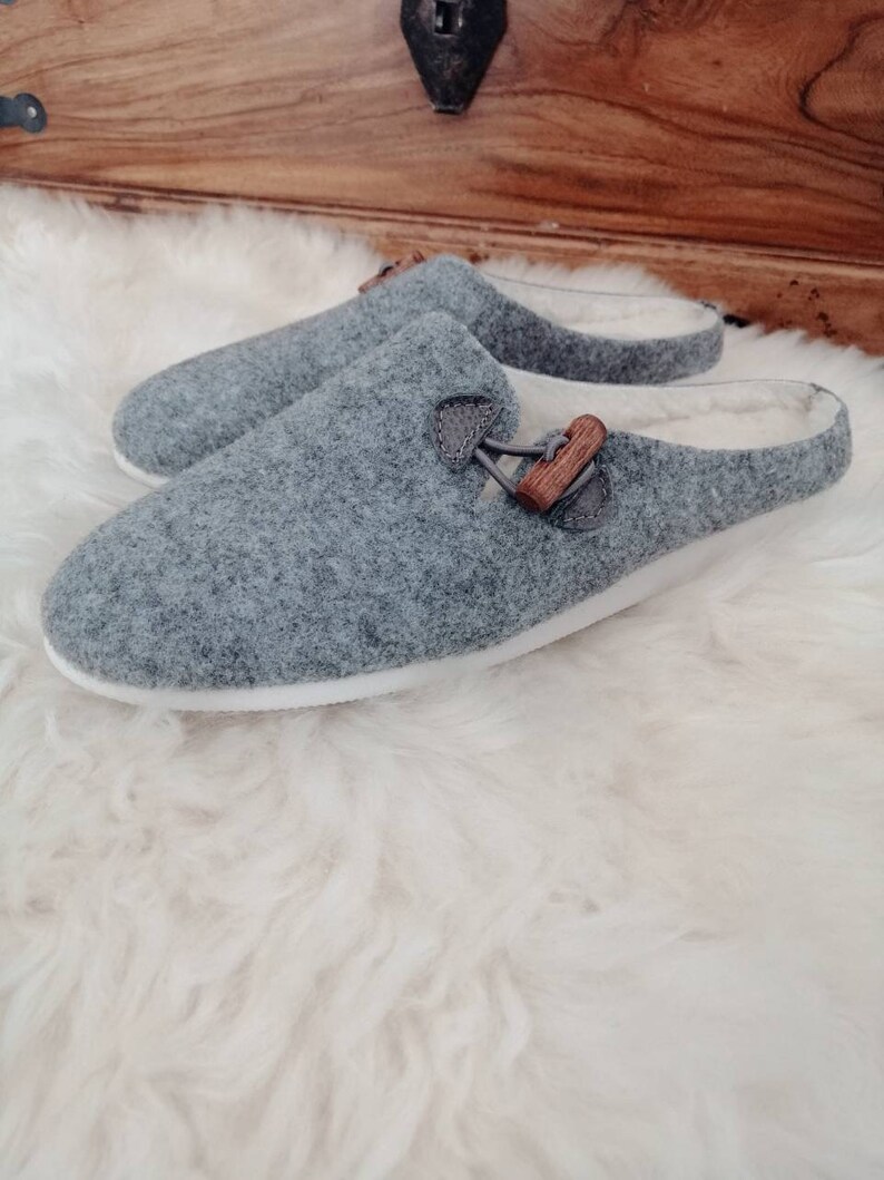 May include: A pair of grey wool slippers with a wooden buckle detail. The slippers have a white sole and a soft, fuzzy lining.