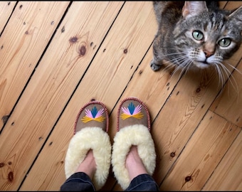 May include: A pair of brown suede slippers with white sheepskin lining and colourful embroidered designs. The slippers are on a wooden floor with a tabby cat looking up at the camera.