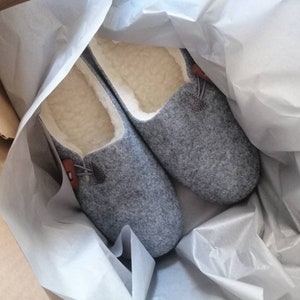 May include: A pair of grey wool slippers with a brown leather accent and a white fleece lining. The slippers are in a white tissue paper-lined box.