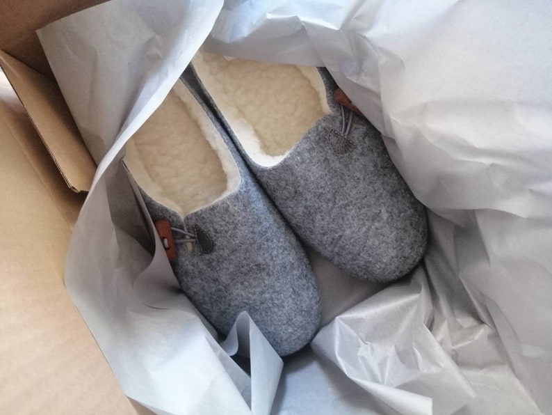May include: A pair of grey wool slippers with a brown leather accent and a white fleece lining. The slippers are in a white tissue paper-lined box.