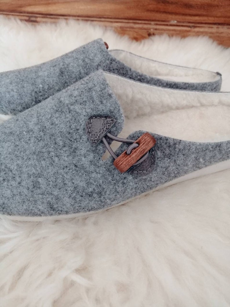 May include: A pair of grey felt slippers with a wooden button detail. The slippers have a white fleece lining.