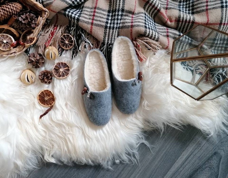 May include: A pair of grey wool slippers with a white fleece lining. The slippers have a brown leather accent on the top. The slippers are sitting on a white fluffy rug.