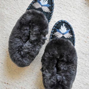 May include: A pair of dark grey, plush slippers. The slippers have a dark blue suede upper with embroidered floral designs in white, light blue, and grey. The edges are trimmed with a braided blue cord.