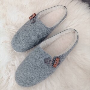 May include: A pair of grey wool slippers with a wooden button detail. The slippers have a white fleece lining.