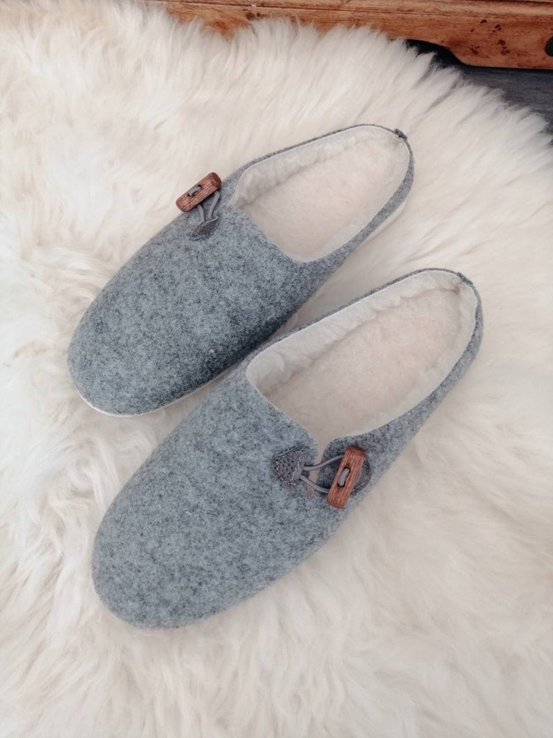 May include: A pair of grey wool slippers with a wooden button detail. The slippers have a white fleece lining.