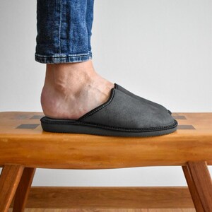 May include: A close-up of a gray slipper with black trim. The slipper is on a wooden bench. The person is wearing blue jeans. The slipper appears to be made of a soft material.
