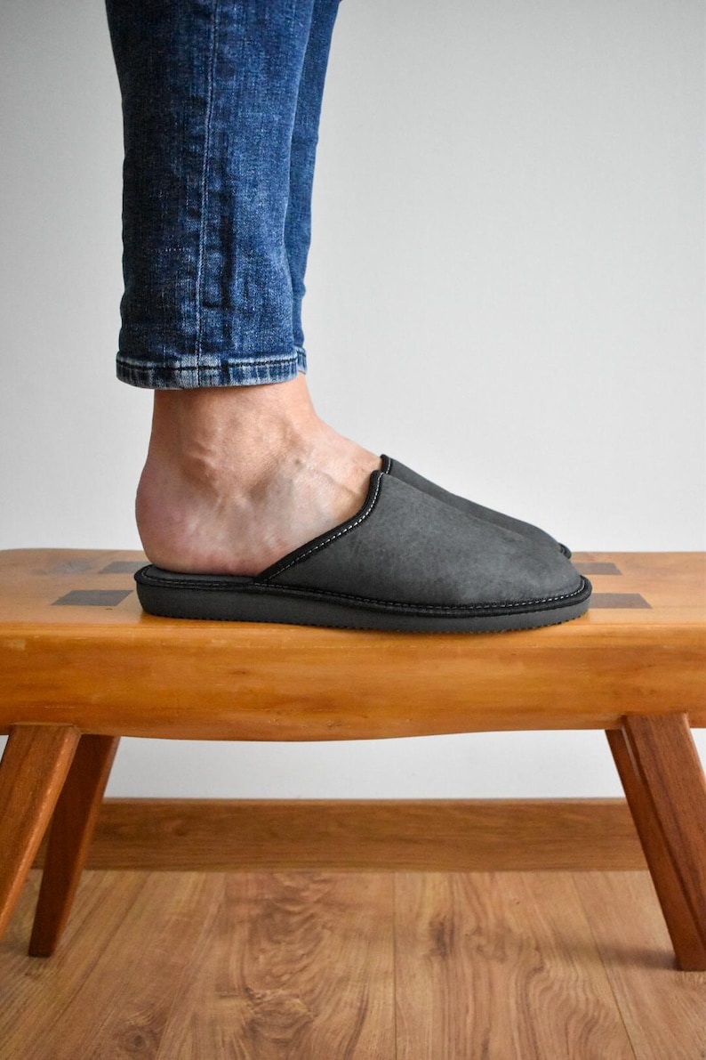 May include: A close-up of a gray slipper with black trim. The slipper is on a wooden bench. The person is wearing blue jeans. The slipper appears to be made of a soft material.
