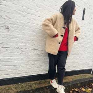 May include: A woman wearing a cream-colored shearling jacket with black trim, a red sweater, black pants, and white high-top sneakers with red accents.