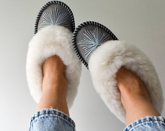 May include: A pair of gray sheepskin slippers with white fur trim and blue embroidery. The slippers are worn by a person wearing blue jeans.
