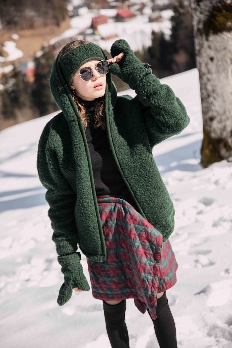 May include: A woman wearing a green fleece jacket with a hood, a black turtleneck, and a red and green plaid skirt. She is wearing sunglasses and a headband.