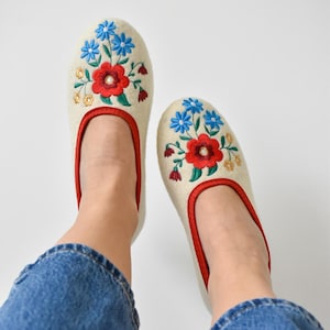 Puede incluir: Un par de zapatillas color crema con ribete rojo y diseños florales bordados en rojo, azul y amarillo. Las zapatillas las lleva una persona con vaqueros azules. Calzado cómodo.