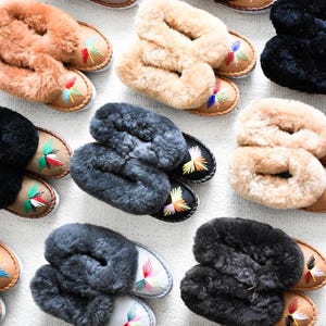 May include: Overhead shot of multiple pairs of cozy slippers in various colors. The slippers have a soft, fluffy upper and a leather sole. Each slipper features colorful embroidered accents on the toe.