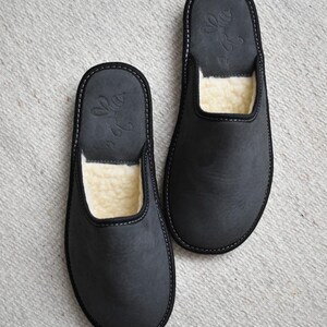 May include: A pair of dark gray leather slippers with a soft, cream-colored interior. The slippers have a simple design with a stitched border and a decorative emblem on the top.