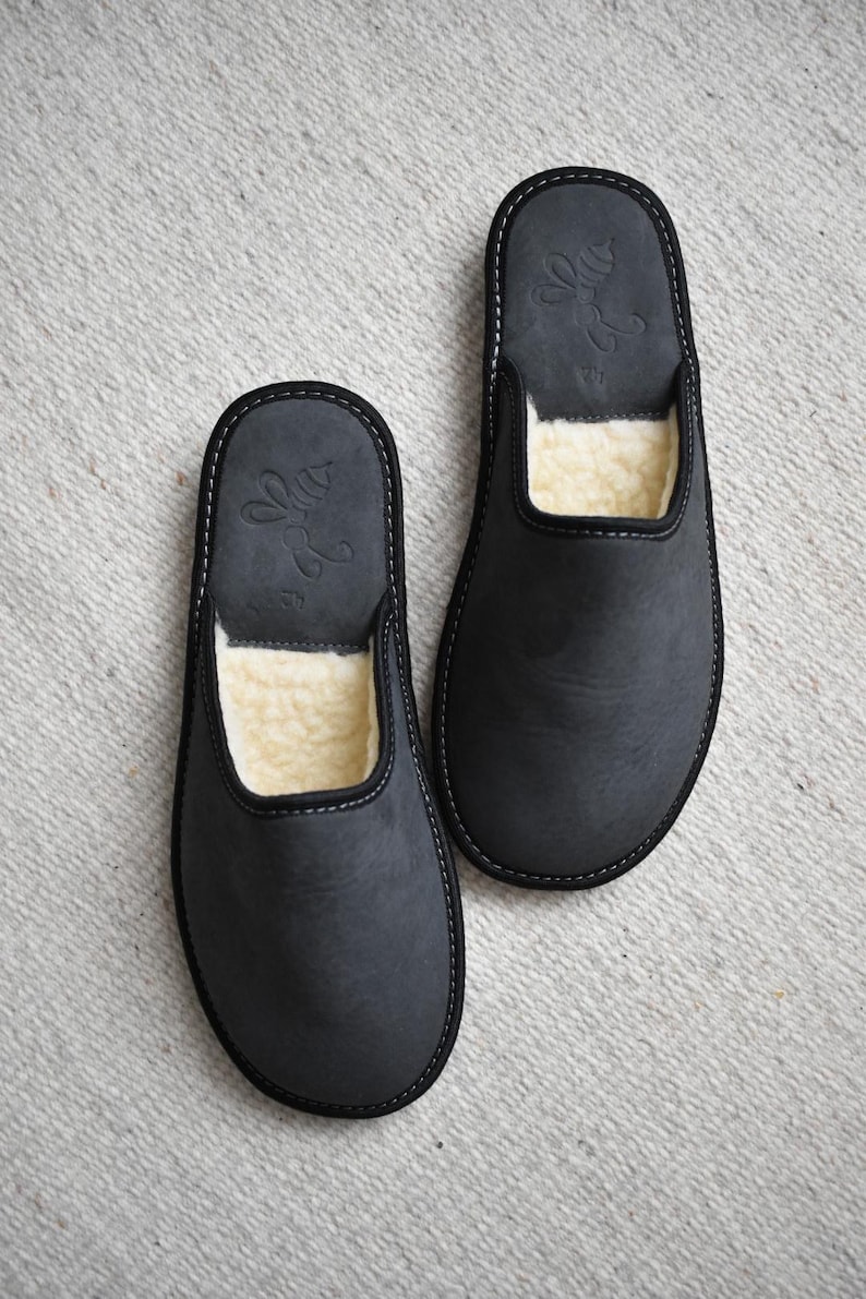 May include: A pair of dark gray leather slippers with a soft, cream-colored interior. The slippers have a simple design with a stitched border and a decorative emblem on the top.