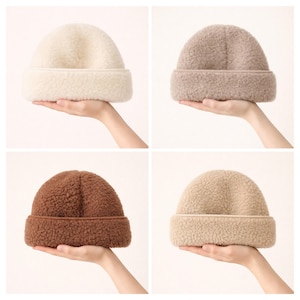 May include: Four fleece beanies in cream, taupe, brown, and tan are shown. Each beanie has a folded brim and a soft, textured finish. The beanies are held in place by a hand. The hats are a winter accessory.