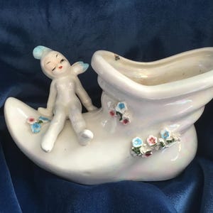 May include: A white ceramic planter shaped like a shoe with a small fairy figurine sitting on top. The planter is decorated with blue and pink flowers.