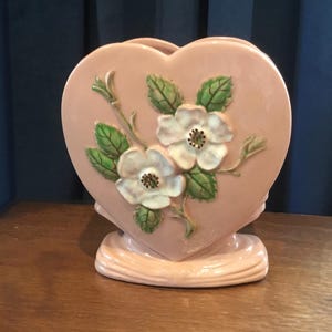 Puede incluir: Un jarrón de cerámica rosa en forma de corazón con un diseño floral en relieve. El jarrón presenta dos flores blancas con hojas y tallos verdes.