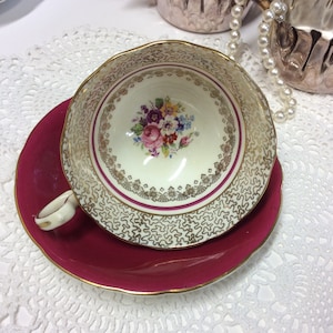 May include: A vintage teacup and saucer set featuring a floral design and gold trim. The teacup has a white background with a pink, yellow, and purple floral pattern. The saucer is a deep burgundy red.