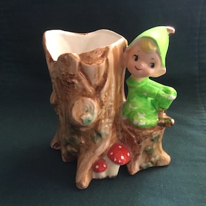Könnte beinhalten: Ein Keramik-Pflanztopf in Form eines Baumstumpfes mit einer grünen Elfenfigur darauf. Der Elf trägt einen grünen Hut und ein grün-weiß gemustertes Outfit. Am Fuß des Baumstumpfes befinden sich rote und weiße Pilze.