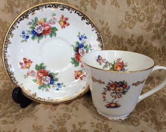 May include: A white teacup and saucer with a gold rim and floral pattern. The flowers are red, orange, yellow, blue, and purple.