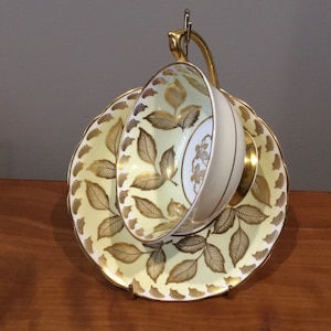 Könnte beinhalten: Ein Vintage-Tee-Set mit Tasse und Untertasse. Die hellgelbe Untertasse und die Innenseite der Tasse weisen ein goldenes Blattmuster auf. Die Tasse ist weiß mit goldenem Rand und floralem Design. Das Set wird auf einem goldfarbenen Metallständer präsentiert.
