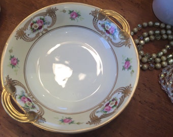 May include: A white porcelain bowl with gold trim and floral designs. The bowl has two gold handles and is perfect for serving snacks or desserts.
