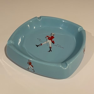 May include: A light blue ceramic ashtray with a red and white illustration of a man in a red jacket and white pants. The text "Born 1820 - Still Going Strong" is printed on the ashtray.