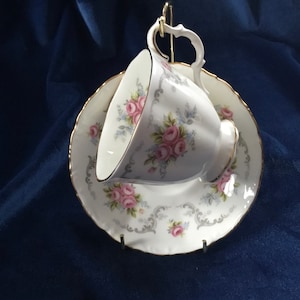 Könnte beinhalten: Eine weiße Teetasse und Untertasse mit einem zarten Blumenmuster mit rosa Rosen und grauen Schnörkeln. Tasse und Untertasse haben einen goldenen Rand.