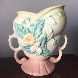 Puede incluir: Un jarrón de cerámica vintage con cuerpo en forma de corazón y dos asas. El jarrón está decorado con un diseño floral en tonos verdes, amarillos y rosas.