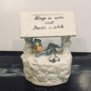Pode incluir: Um poço dos desejos de cerâmica branca com uma figura de elfo azul deitada dentro. O poço tem uma placa que diz "Drop a coin and make a wish".