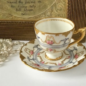 May include: A delicate teacup and saucer set with a floral and gold design. The cup and saucer are white with a blue and gold band around the rim. The design features pink roses and gold accents.